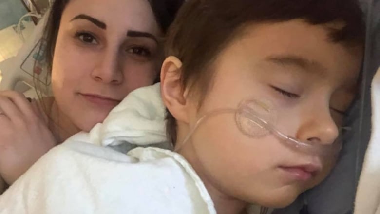 A child lies sleeping in a hospital bed, with an oxygen tube attached to his nose. His mother lies beside him, looking at the camera.