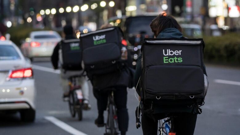 Men ride bikes in the road wearing Uber Eats delivery bags on their backs, 