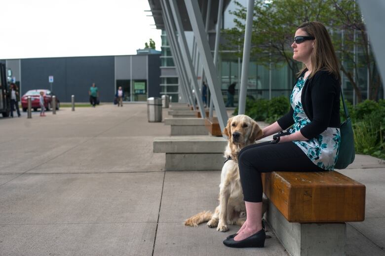 A woman wearing dark glasses sits on a bench petting a white dog.