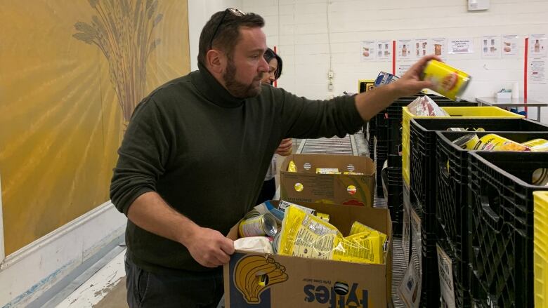 A man in a warehouse stands in front of a row of bins and boxes. He reaches toward one with a can of food in his hand.