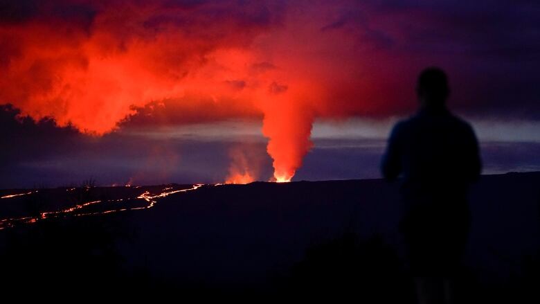 A person is silouetted in the foreground as a cloud of smoke illuminated by burning lava spews upwards. 