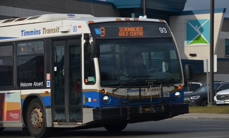 A Timmins transit bus reading 'Schumacher/Gold Centre' turns a corner in front of a parking lot. 