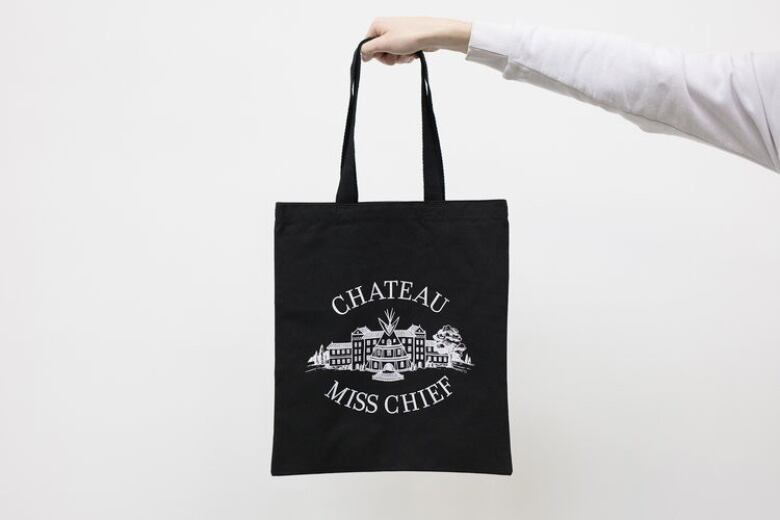 Photo of an outstretched arm holding a black canvas tote. The tote's printed design depicts a Victorian home with a tipi incorporated into the facade. It reads 