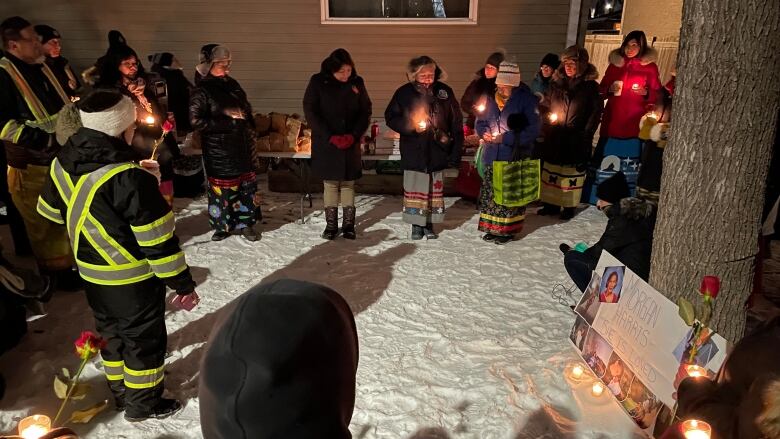 People in a circle hold candles and roses as they stand on snowy ground. Nearby, a poster with photos of a young woman sits propped up on a tree.