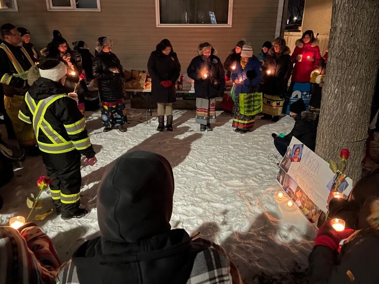 People in a circle hold candles and roses as they stand on snowy ground. Nearby, a poster with photos of a young woman sits propped up on a tree.