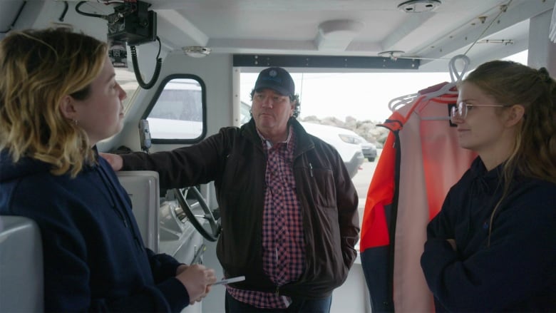 A man speaks to two women in the wheelhouse of a boat.