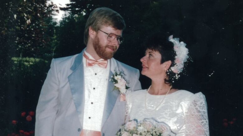 A man in a suit and bowtie gazes into the eyes of a woman wearing a white dress and carrying flowers. 