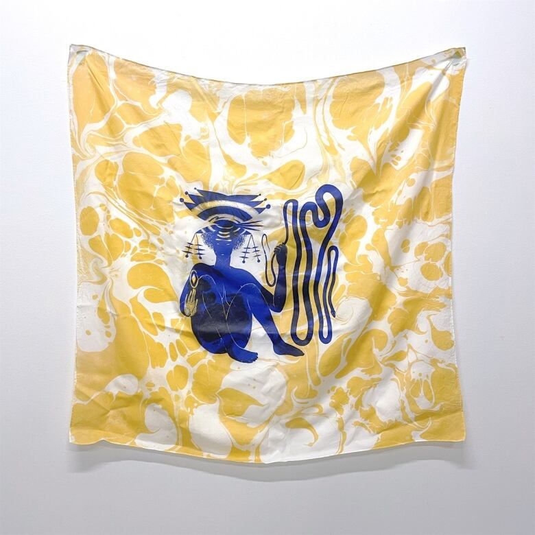 Photo of a printed silk scarf hanging on a white wall. Its background is a yellow abstract pattern. At the centre is a seated figure in blue.