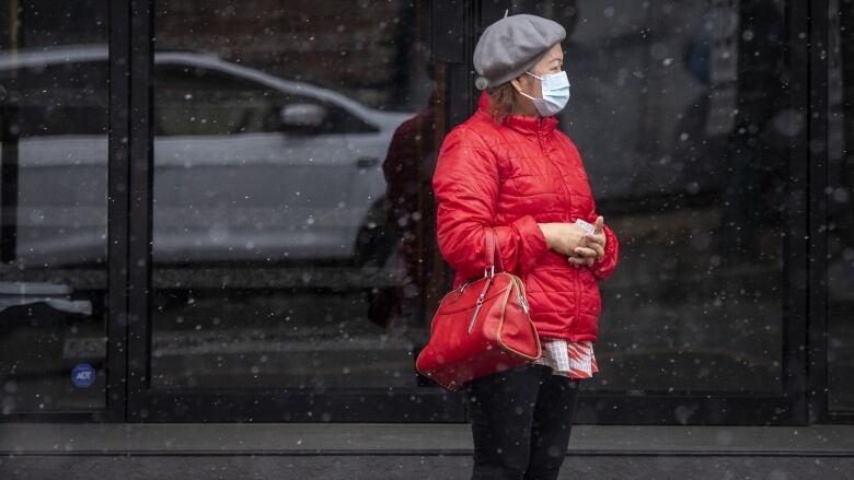 A woman wears a medical masks while standing on the street during snowfall.