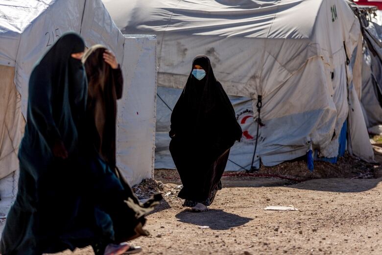 Women dressed in all black walk in front of white tents in a desert.