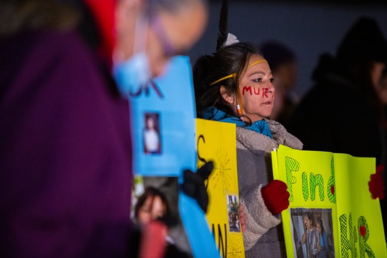 A woman wearing winter clothse stands among a crowd of others outsides, holding a sign reading 