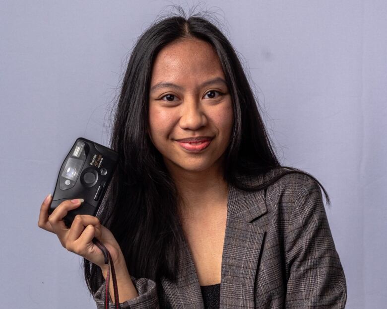 A young woman holding a camera in her right hand poses and smiles at the camera.