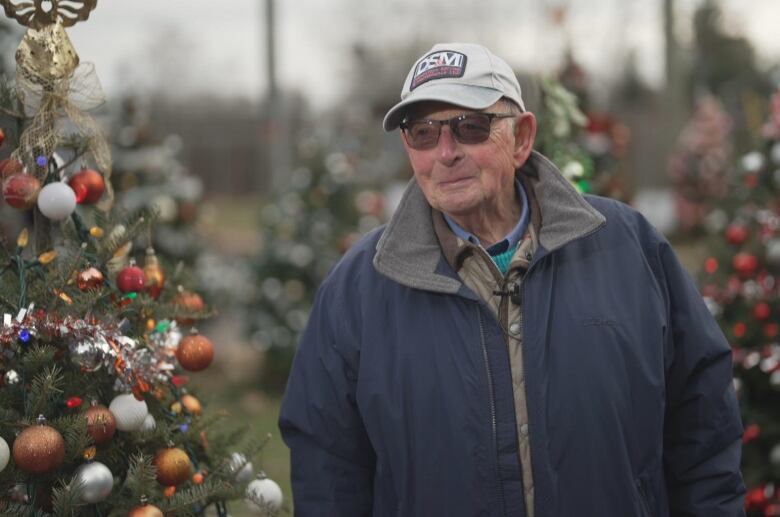 An older man with a jacket and ball cap stands in front of two decorated Christmas trees. 