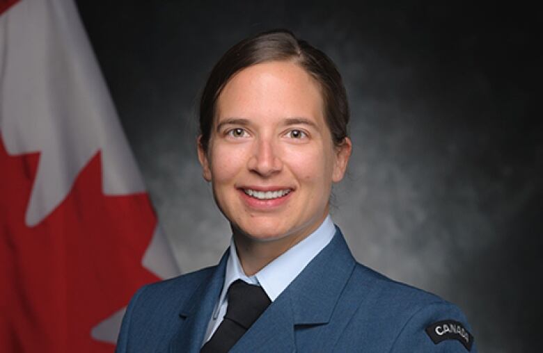 A smiling, dark-haired woman in a blue uniform sits with her hands folded on her lap, in front of the Canadian flag.