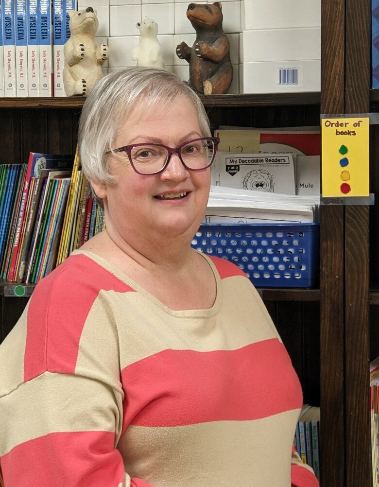 A woman with grey hair and glasses stands in front of a wooden bookcase.