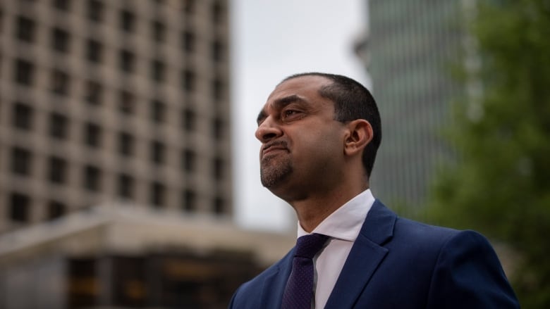 A South Asian man stares off to the left while being flanked by tall buildings in downtown Vancouver.