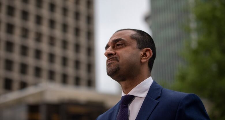 A South Asian man stares off to the left while being flanked by tall buildings in downtown Vancouver.