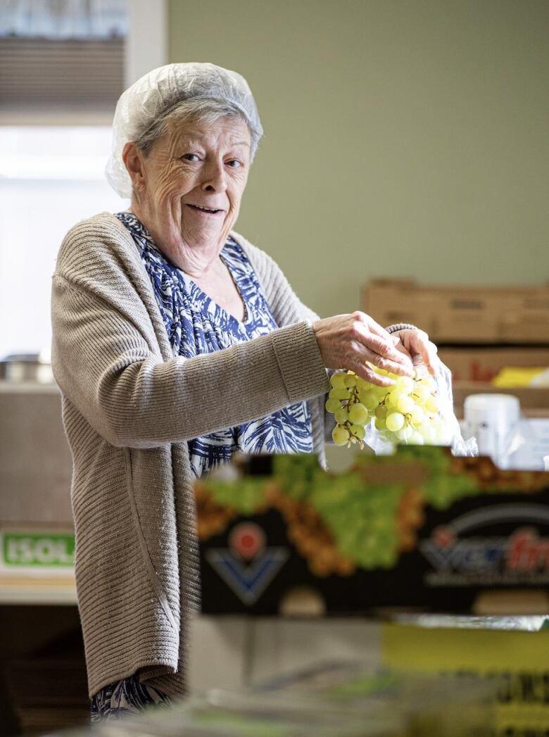 Oder woman, grey hair, with hair net, holds a bunch of green grapes and she prepares an order at the food bank.