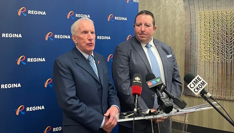 Two men in suits stand in front of microphones at a podium with a backdrop featuring the City of Regina logo behind them.