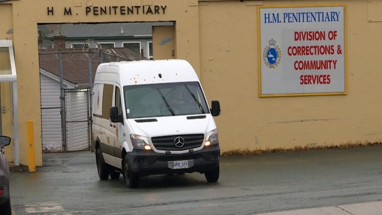 A white van is driving from a yellow stone building. 