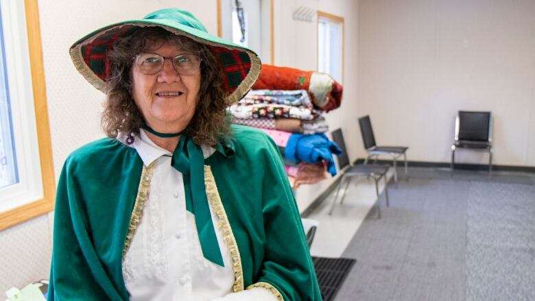 A woman dressed in Christmas clothes stands in front a room with chairs and blankets for unhoused people.