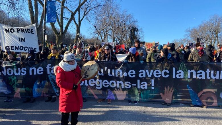 a woman in a red coat and white scarf playing a drum in front of thousands of protesters on the streets of Montreal.