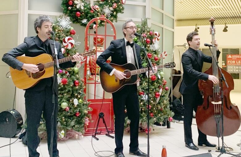 Three men wearing black tuxedos play guitar and bass in front of Christmas decorations