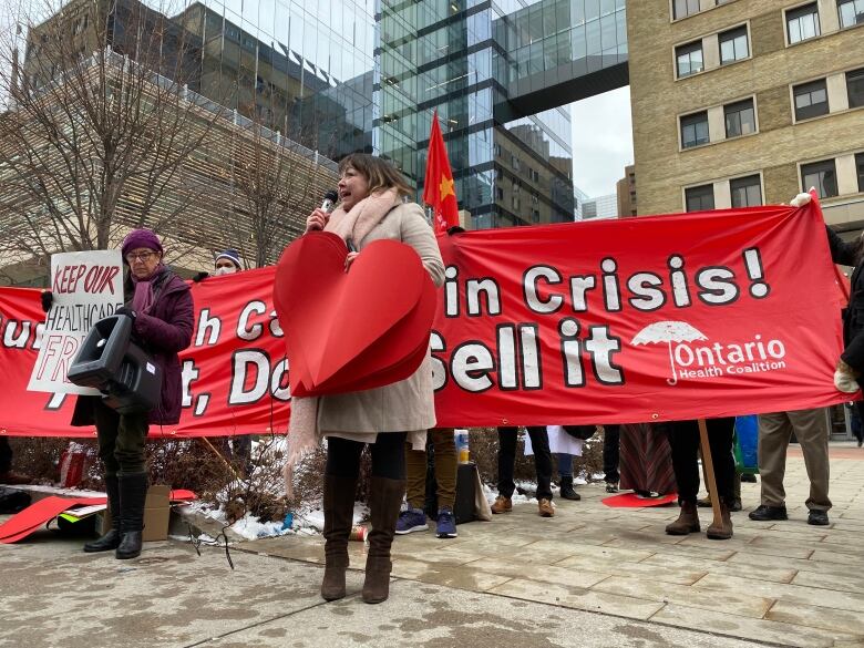 A woman in a beige coat holding a large red hart in front of a sign reading Ontario Health Coalition speaks into a microphone