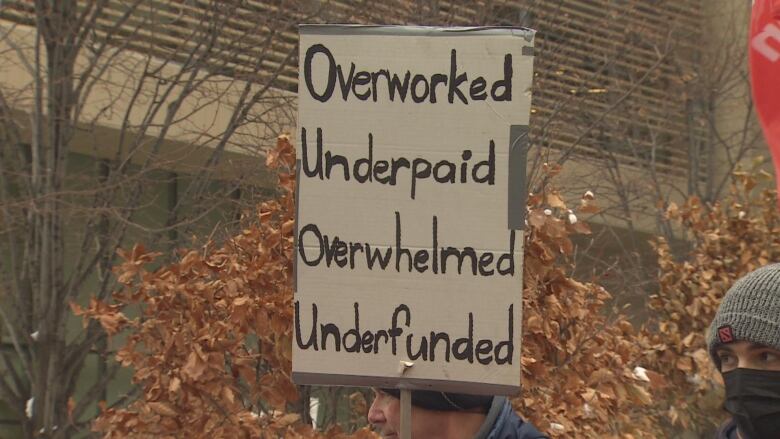 A sign that reads Overworked, underpaid, overwhelmed, overfunded. held in front of trees with falling leavs