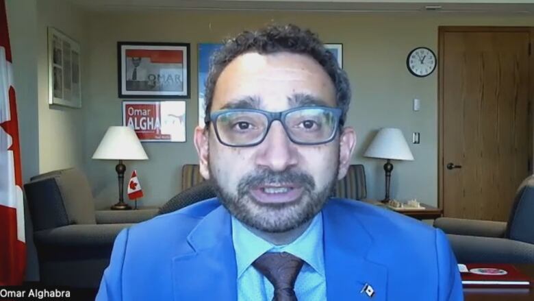 A man with glasses and a goatee speaks on Zoom in an office room with Canadian flags on the wall and a sign that says Omar Alghabra in the background.