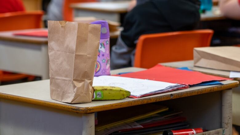 A brown bag for lunch sits on a child's desk in a classroom. 