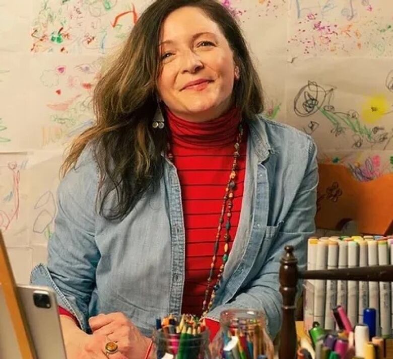 A smiling woman sits in front of jars filled with coloured pencils and markers.