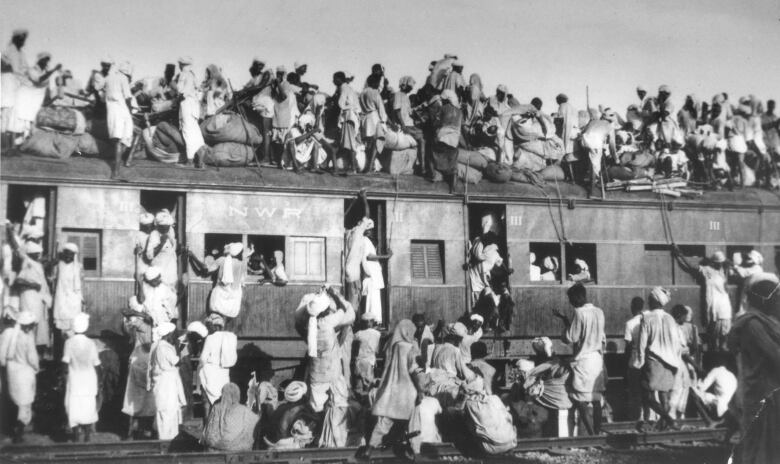 Hundreds of Muslim refugees are on top and in an overcrowded train, also outside on the platform. There are people also coming out of the doors and windows. This is a black and white image.