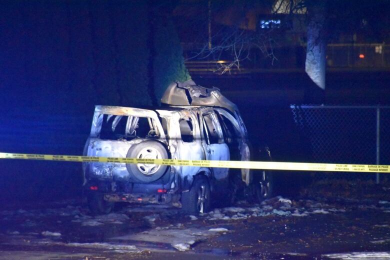 A burnt SUV is pictured in a parking lot behind yellow police tape near a sports field at night.