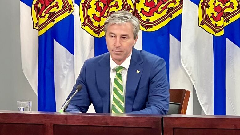 Nova Scotia Premier Tim Houston is seen wearing a navy blue suit and a green stripped tie on sitting at a mahogany desk. Behind him are four Nova Scotia flags.