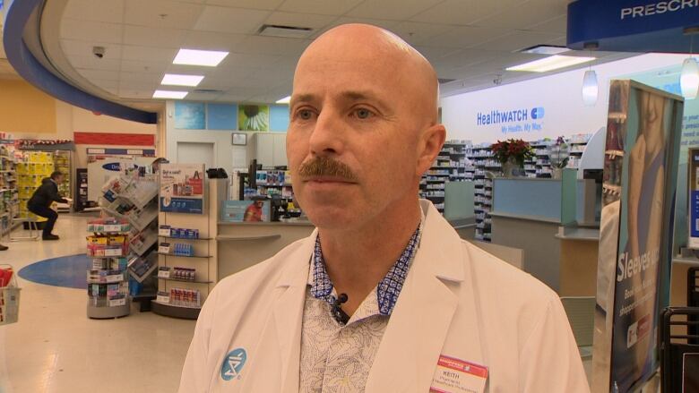 A man wearing a white lab coat standing in a pharmacy. 