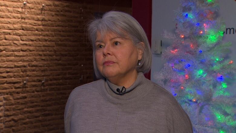 A woman wearing a grey sweater standing in front of a brick wall. A Christmas tree is in the background. 