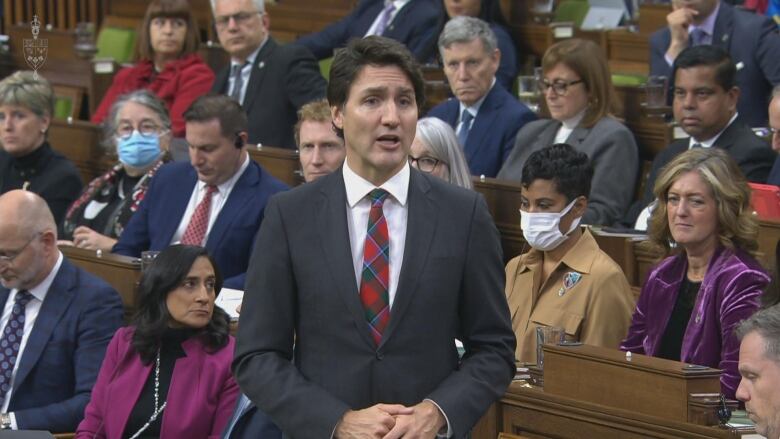 A man with short dark hair in a grey suit and a red plaid tie stands in front of a group of people sitting at desks.