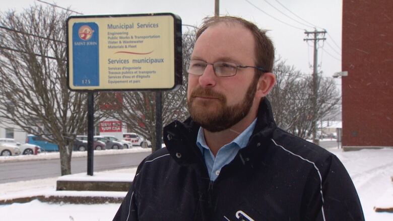 A man stands outside in the snow, we see him from shoulders up. He is wearing glasses, a blue button down, a black winter coat and has a beard. 