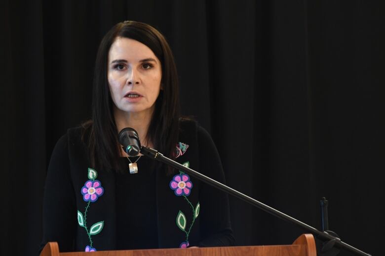 A woman with dark hair past her shoulders and wearing a black shirt with pink-and-blue beaded flowers speaks into a microphone.