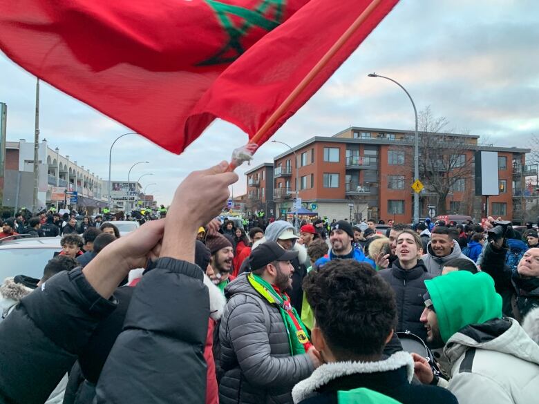 Morocco fans cheer and drum on Jean-Talon Avenue. A man waves a Moroccan flag overhead.