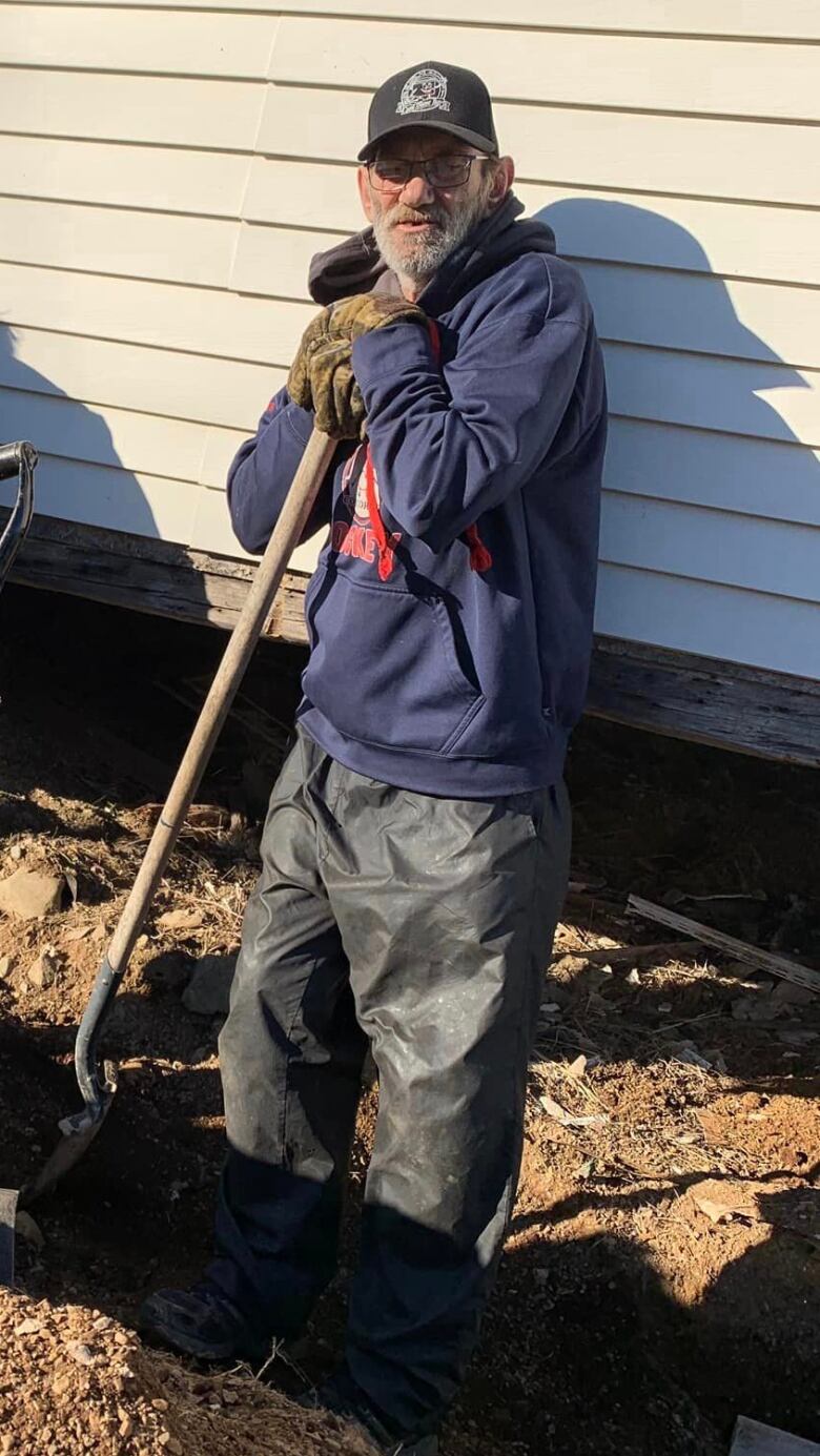 A man stands in a hole wearing work gloves, leaning against a shovel. 