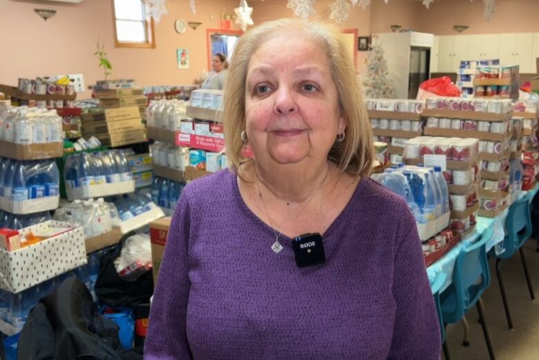 A woman in a purple knit sweater stands in front of a room full of Christmas food hampers. 