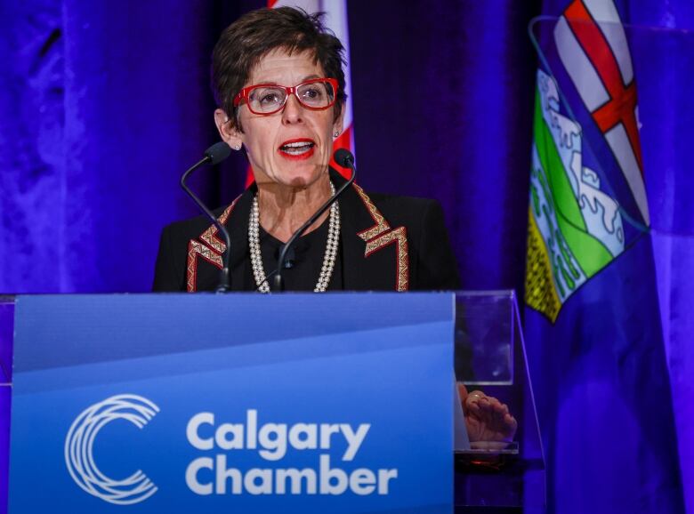 A woman wearing bright red glasses stands behind a podium that has Calgary Chamber written across it. Behind her is a blue backdrop and an Alberta flag. 