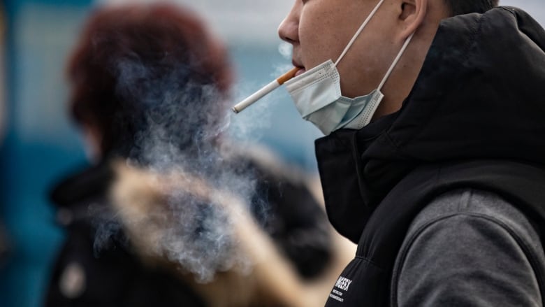 A man smokes a cigarette with his blue surgical mask pulled down to his chin.