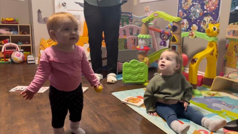 Two children aged around 18 months stand and sit on a playmat. 