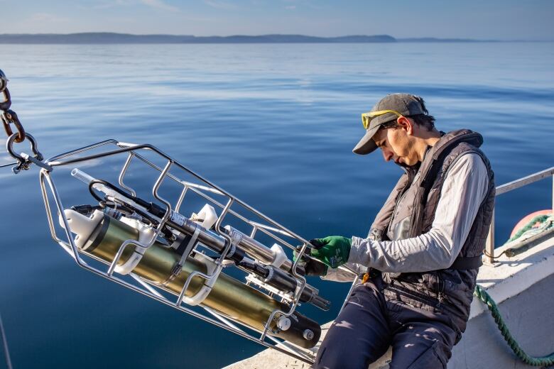 Man in a lifejacket leans on the side of a boat, holding a large piece of scientific equipment.