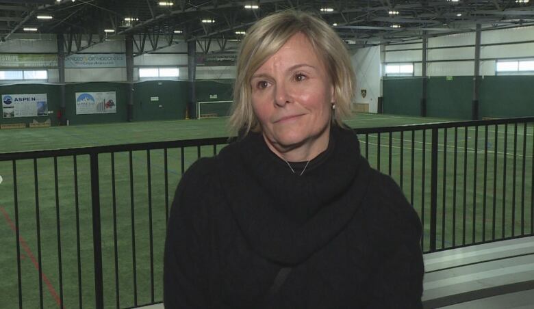 A blond woman wearing a black sweater stands before an indoor soccer pitch. 