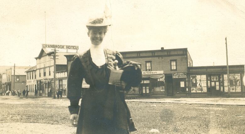 A woman in dress and with a hat stands in front of Cranbrook Hotel and other buildings on the main drag.