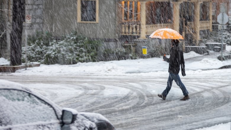 A pedestrian crosses a residential street in the West End of Vancouver, holding an umbrella to guard against snow.